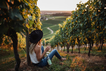 Junge blonde Frau, die sich in den Weinbergen in der Sommersaison entspannt. Mädchen, das nahe der Flasche sitzt und Glasweißwein hält. Outdoor-Bauer im Landhausstil