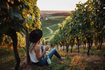 Poster Jonge blonde vrouw ontspannen in de wijngaarden in het zomerseizoen. Meisje zit in de buurt van de fles en houdt glas witte wijn vast. Outdoor boer landelijke stijl © szaboerwin