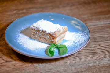 Layered cake with cream Napoleon, vanilla slice with mint and sugar powder served on blue plate on wooden table.