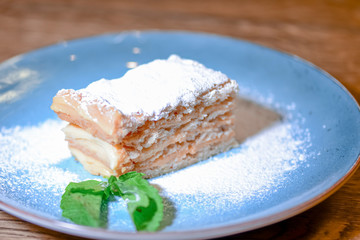 Layered cake with cream Napoleon, vanilla slice with mint and sugar powder served on blue plate on wooden table.