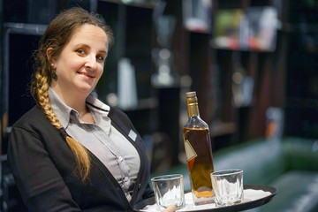Waitress carries a bottle of whiskey with glass on a tray to the client of the hotel bar. The concept of service.