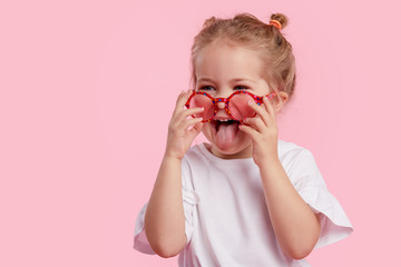 Portrait of surprised cute little toddler girl in the heart shape sunglasses. Child with open mouth having fun isolated over pink background. Looking at camera. Wow funny face