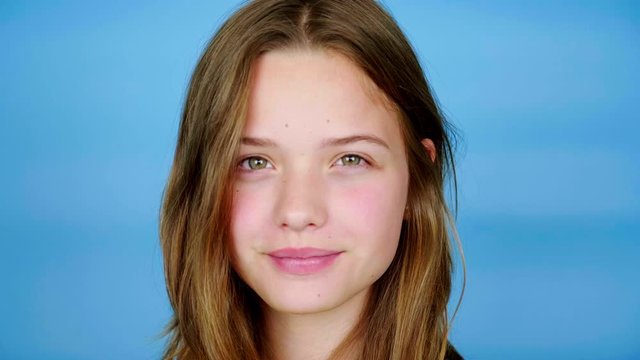 Close-up of teen girl in a yellow t-shirt who looking at camera, winks and smile. Blue background with copy space. Teenager emotions. 4k footage