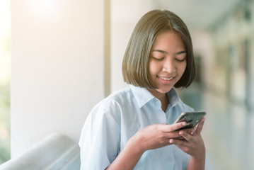 Fototapeta na wymiar A smiling Asian female high school student in white uniform is sitting and enjoying social media on her smartphone in the school.