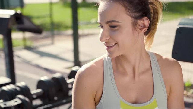 Smiling sporty woman takes off wireless earphones while sitting on bench in outdoor gym