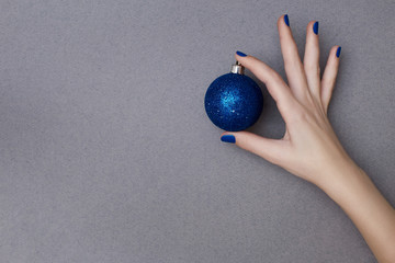 Female hand with manicure in trendy classic blue color holding sparkling blue christmas decorative ball on the grey background