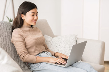 Pretty asian girl using laptop, relaxing on sofa at home