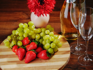 Fresh green grapes and red strawberry on a wooden board, Two champagne flutes  and a bottle of champagne without labell, flower in the background, close up. Selective focus.