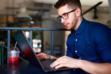 Thoughtful young attractive hipster freelancer working remotely on laptop in loft cafe. Businessman browsing internet at coffeshop. Blogger posts to social media using internet. Telecommuting theme.