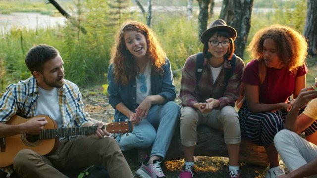 Multiracial group of tourists friends is having fun in forest playing the guitar and singing songs in the woods. Friendship, music and active lifestyle concept.