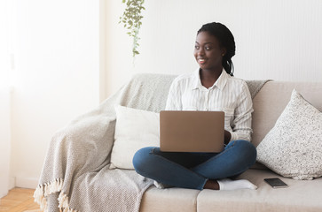 Afro woman sitting on couch with laptop, working online from home