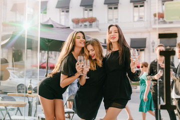 Outdoor portrait of shapely girl in black outfit hanging out with friends around city in morning. Photo of three pretty ladies dancing and enjoying wine on the street with crowd on background.