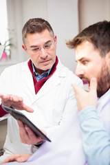 Serious middle-aged dentist or doctor consulting with worried young male patient, showing therapy details on a tablet. Healthcare and medicine concept.