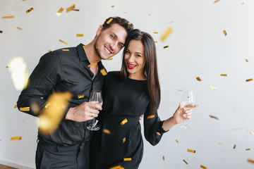 Handsome brunette man embracing his girlfriend and looking at golden confetti at party. Dark-haired lady in elegant dress celebrating something with husband and drinking champagne.