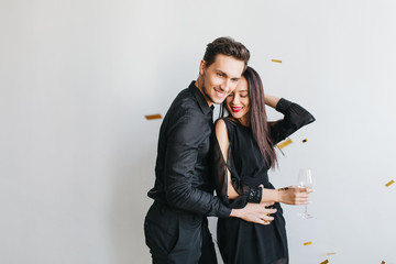 Graceful dark-haired woman leaning to her husband with eyes closed and dreamy smile isolated on white background. Indoor photo of confident man in black outfit embracing cute brunette girl on party.