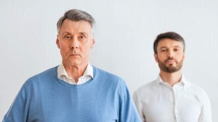 Serious Senior Man And Middle-Aged Son Standing Against White Wall