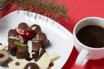Chocolate fudge garnished with chocolate, mint and cranberries. Lies on a white plate, on a white background. Nearby a cup of coffee and a branch of spruce.
