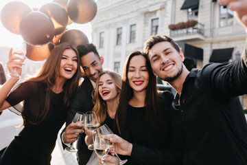 Pretty lady with gently smile posing beside husband while he making selfie after street fest. Outdoor portrait of romantic long-haired woman with party balloons enjoying walk with friends.
