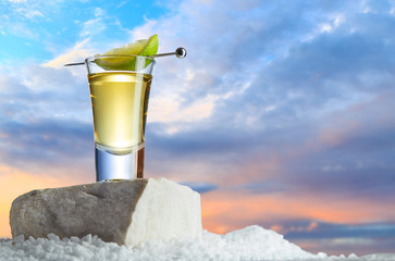 Gold tequila in a glass with slice of lime on a skewer stands on a stone against a beautiful sunset...