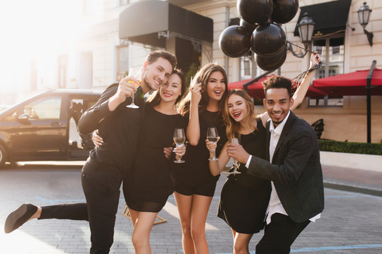 Pleased international students with helium balloons celebrating graduation. Outdoor portrait of happy friends having party on the street and drinking champagne.