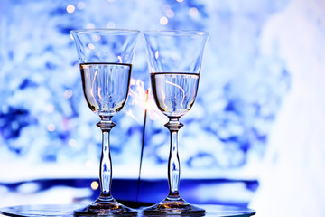 Two glasses with sparkling champagne wine and a garland with lights on the background of frosty patterns of branches in the snow. The winter mood of winter holidays.
