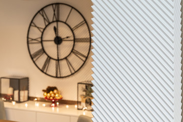 Blurred Christmas decoration with glass ornaments, lights and big modern wall clock on the white striped wall.