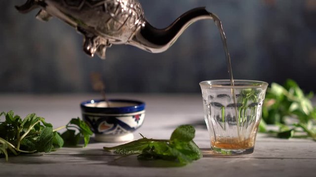 Wide shot of hand picking up an ornate teapot and pours tea into a glass with Moroccan bowl and mint around.