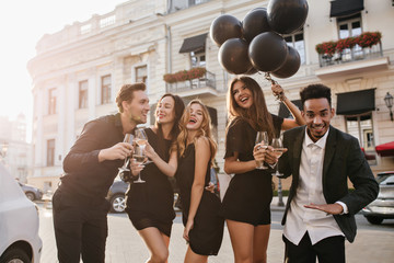 Slim blonde woman with lightly-tanned skin dancing during outdoor photoshot with friends. Black man with african hairstyle waving hand, drinking wine on the street.
