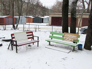 Two benches in the snow stand in the courtyard of an apartment building. Garages for cars.