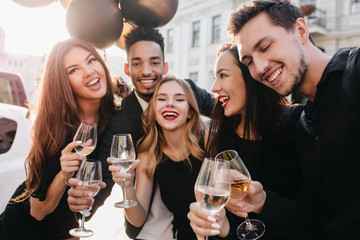 Adorable long-haired lady with nude make-up laughing during outdoor photoshoot with friends. Smiling african male model standing near party balloons on the street in morning.