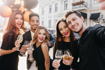 European guy with cute smile making selfie during street fest in sunny day. Adorable caucasian woman waving party balloons while holding wineglass on blur city background.
