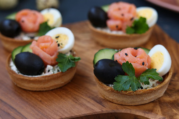 Holiday tartlets with cheese, salmon, black olives, quail eggs and cucumbers on wooden board on dark background, horizontal orientation