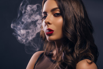 Glamour caucasian brunette woman in evening black dress on a neutral background. She pose sensually, blowing smoke from her mouth. Retro fashion mood.