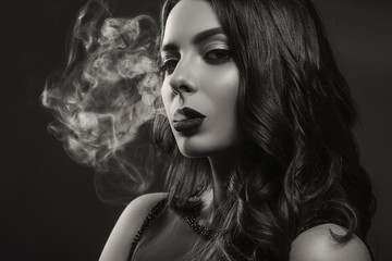 Glamour caucasian brunette woman in evening black dress on a neutral background. She pose sensually, blowing smoke from her mouth. Retro fashion mood.
