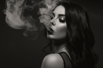 Glamour caucasian brunette woman in evening black dress on a neutral background. She pose sensually, blowing smoke from her mouth. Retro fashion mood.