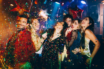 Group of friends at club having fun. New year's party with confetti and sparklers  - obrazy, fototapety, plakaty