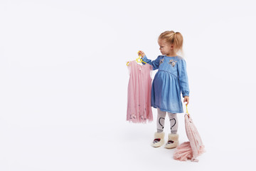 Little child girl chooses outfits for the holiday. Holds hangers with dresses in his hands. White studio background