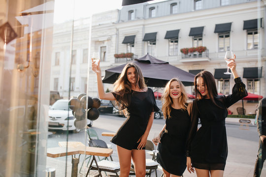 Brunette Girl In Black Dress With Long Sleeves Raising Glass Of Wine And Laughing On Urban Background. Glad Ladies Celebrating Vacation During Travel Around The World.