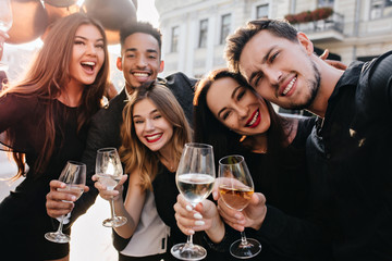 Close-up portrait of laughing young people enjoying summer on the street and drinking champagne. Enthusiastic blonde woman holding wineglass and posing with big sincere smile o blur city background.