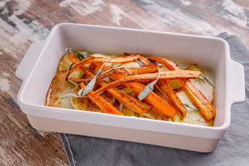 Closeup baked sliced potatoes and carrots with red onions and herbs: sage, rosemary and thyme in a ceramic baking dish