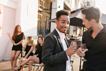 Beautiful girls with long legs sitting at street cafe while guys on foreground clinking glasses. Funny african man in stylish jacket talking with friend, while drinking wine.