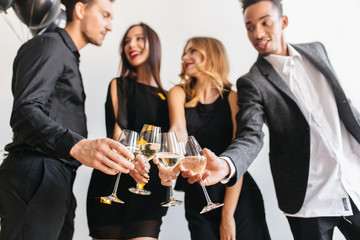 Indoor portrait of brunette girl clink wineglasses with friends and looking at brunette guy. African man in stylish white shirt having fun at home party and enjoying champagne.