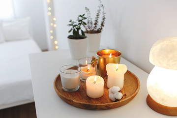 Fototapeta na wymiar Wooden tray with burning candles, night lamp and home plants in pots standing on white table in bedroom. Christmas cozy winter home decor. New year interior decorations. White stylish room.