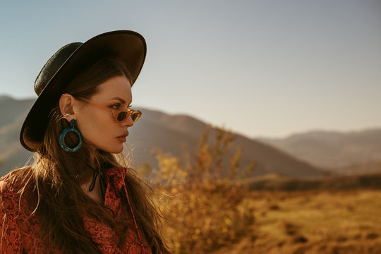 Outdoor close up fashion portrait of young beautiful confident brunette woman wearing stylish black wide brimmed hat, blue hoop earrings, orange dress, posing in mountain landscape. Copy, empty space