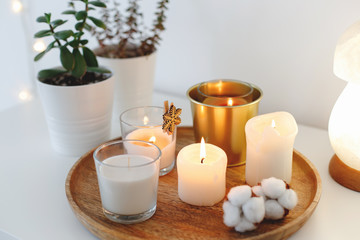 Fototapeta na wymiar Wooden tray with collection of different burning candles, night lamp and home plants in pots standing on white table. Cozy winter home decor. Interior decorations. Stylish composition, close up.