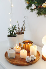 Wooden tray with burning candles, night lamp and home plants in pots standing on white table. Christmas cozy winter home decor. New year interior decorations. Stylish composition.