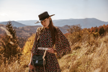 Outdoor fashion portrait of young beautiful confident brunette woman wearing stylish hat, chiffon flower print dress, white belt, with crossbody bag, posing in mountain landscape. Copy, empty space - obrazy, fototapety, plakaty