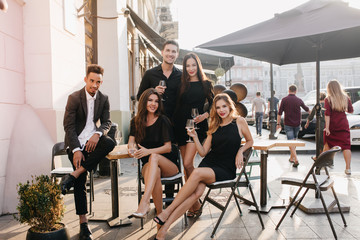 Stylish african man sitting in confident pose and looking at girls in outdoor cafe. Portrait of happy couple posing on the street with friends enjoying meeting.