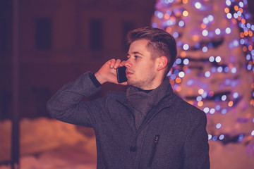 Man talking on the mobile phone while waiting for someone at winter night in decorated city street.