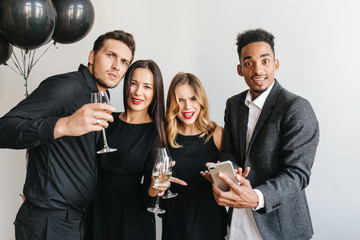 Surprised guy with dark eyes and brown skin showing phone message to blonde girl. Indoor portrait of relaxed friends in black clothes enjoying fest and drinking champagne.